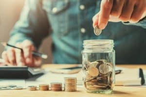 A person in a denim shirt carefully places coins into a glass jar filled with money while using a calculator in the background, symbolizing how financial growth comes from small steps in saving and budgeting.