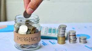 Hand placing a coin into a glass jar labeled "Emergency," surrounded by stacks of coins and financial charts on the table. The image symbolizes building an emergency fund as an essential part of managing a budget effectively.