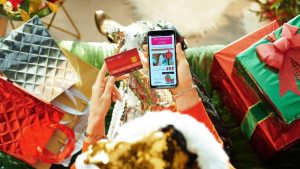 Person holding a credit card and a smartphone displaying an online store while surrounded by holiday gifts and shopping bags. The image highlights the importance of using coupons and cashback to save money during festive purchases.