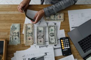 Person organizing stacks of U.S. dollar bills on a desk filled with financial documents, a laptop, and a calculator app on a smartphone. The image illustrates the importance of discipline in achieving financial goals through budgeting and careful money management.