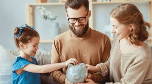 Smiling parents and their daughter putting coins into a piggy bank together at home. The image represents teamwork and the importance of teaching children about managing family finances responsibly.