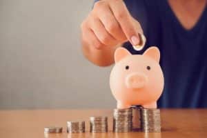 Hand placing a coin into a pink piggy bank surrounded by neatly stacked coins on a table. The image represents staying motivated to save money and building financial security through consistent saving habits.