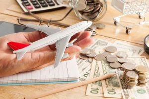Hand holding a toy airplane above a desk with scattered coins, dollar bills, and a notebook, symbolizing budgeting for trips. The image highlights the concept of saving money on travel through financial planning and smart spending.